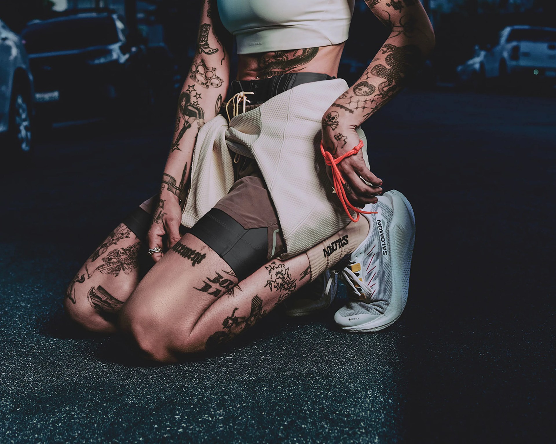 A person kneeling on asphalt at night, wearing a white crop top, brown shorts, and white Salomon sneakers. Their arms, legs, and hands are covered with various black tattoos, and they have a light-colored jacket tied around their waist. A bright orange string is wrapped around their right wrist.