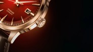 Close-up of a Seiko wristwatch with a red textured dial, gold-tone case, and gold hour markers. The watch features a date window showing the number 6, and the words "PRESAGE AUTOMATIC" are visible on the dial. The crown has an engraved "S" and the watch is paired with a dark leather strap.