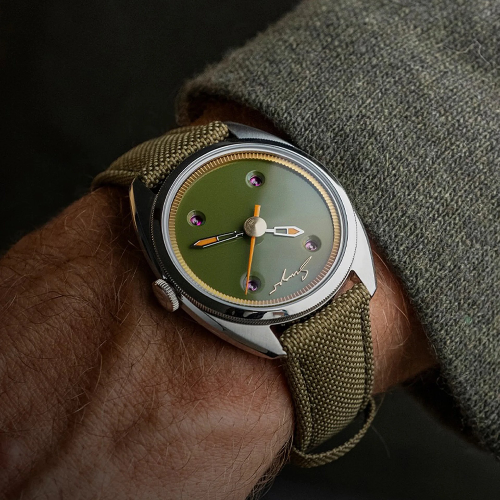 A wristwatch with a green dial featuring four purple jewel-like markers and orange-accented hands. The watch has a polished silver case and a textured olive green fabric strap. The wearer is dressed in a green knit sleeve.