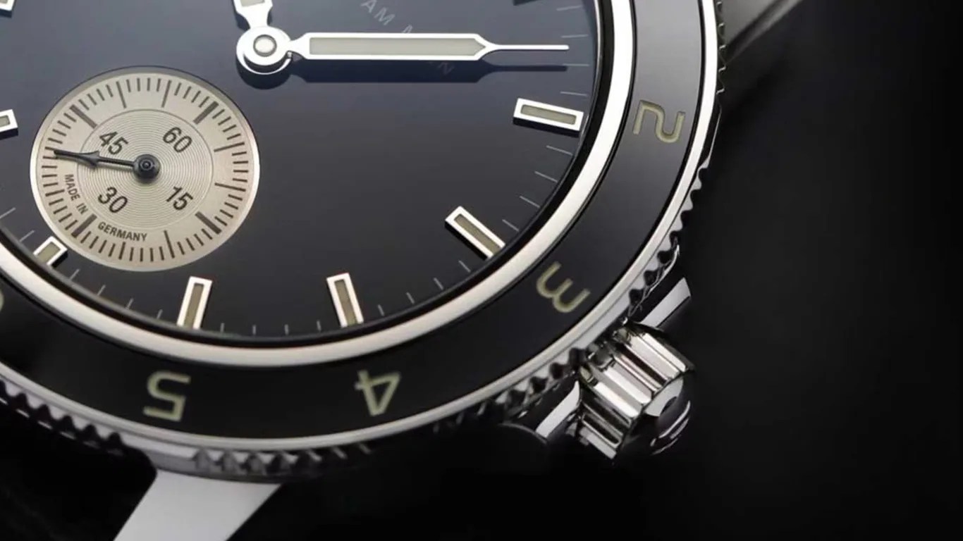 Close-up of a black Sinn wristwatch face with silver hour markers and hands, featuring a small beige subdial marked "Made in Germany" with minute increments. The bezel has numbers 2, 3, 4, and 5 in a light color, and a silver crown is visible on the right side. The watch has a polished metal case and a black background.
