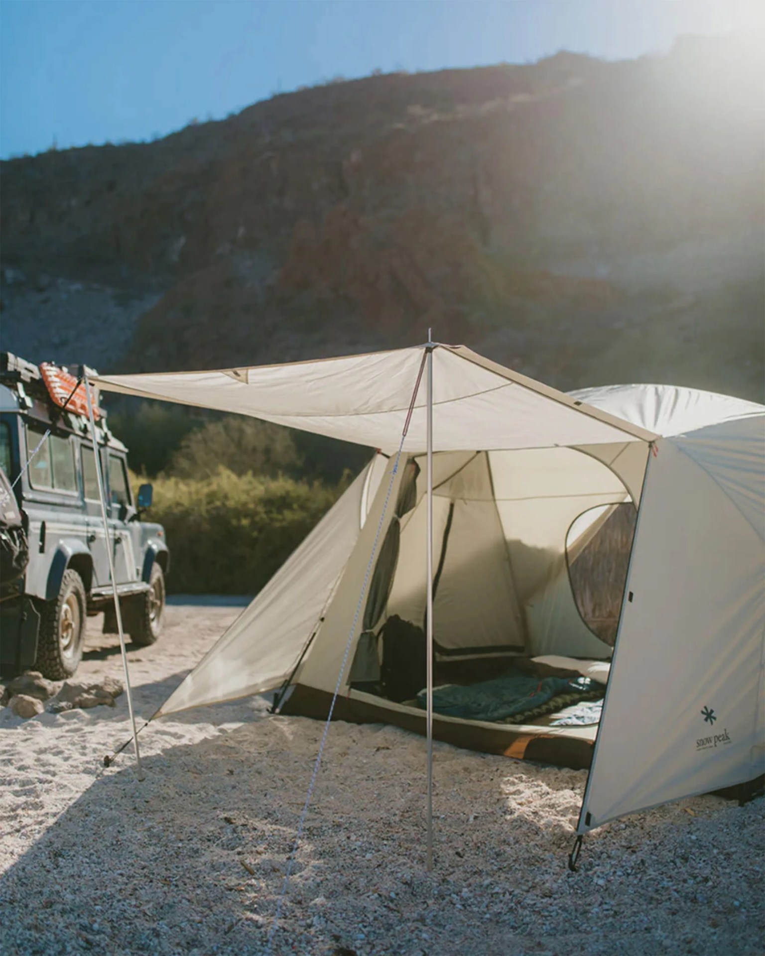 A beige camping tent with an extended canopy is set up on a rocky, sandy ground. Inside the tent, a sleeping bag and some camping gear are visible. To the left of the tent, there is a rugged off-road vehicle equipped with gear on its roof rack. In the background, there are bushes and a large rocky hill under a clear blue sky. The tent has the "snow peak" logo on its side.
