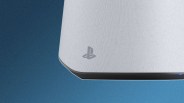 Close-up of the bottom corner of a white PlayStation 5 console, showing the textured surface, the PlayStation logo, and a small blue light on the black base against a blue background.
