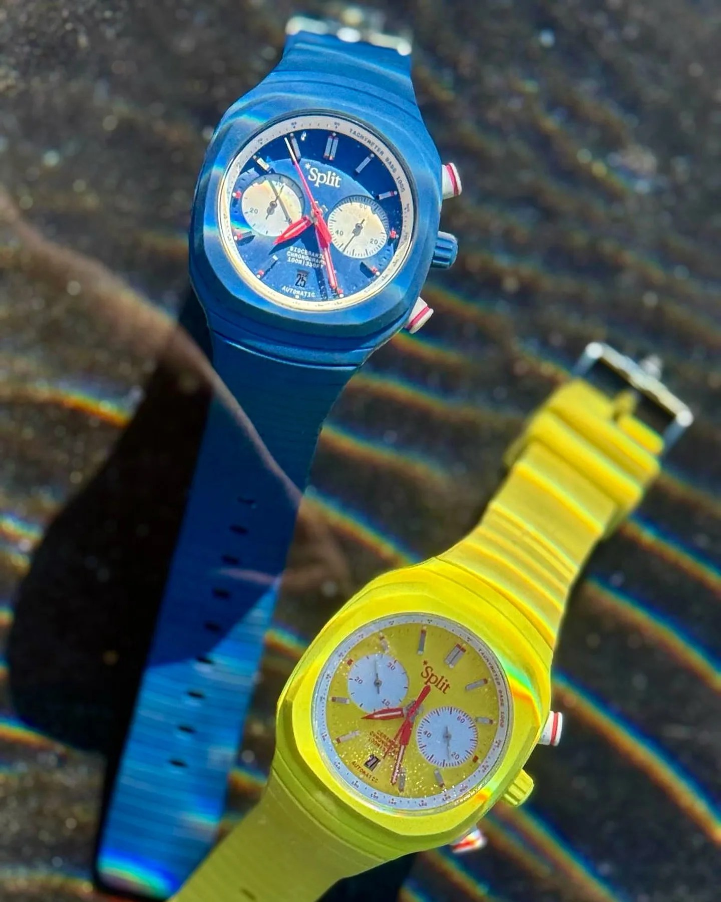 Two wristwatches with rubber straps, one in bright blue and the other in bright yellow. Both watches have round faces with chronograph subdials, red hands, and the brand name "Split" on the dial. The blue watch has a blue dial with white subdials, while the yellow watch has a yellow dial with white subdials. The background shows a textured surface with light reflections creating rainbow-like patterns.