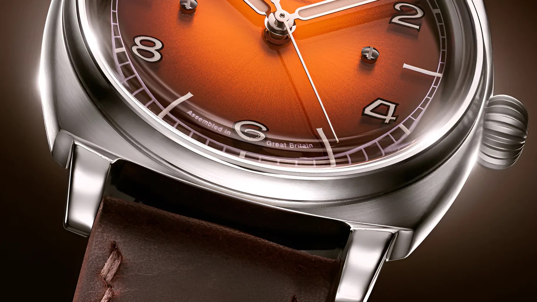 Close-up of a wristwatch with a polished silver case, an orange sunburst dial, and white hour and minute hands. The watch features bold, silver-framed Arabic numerals at 2, 4, 6, and 8 o'clock, and a brown leather strap with visible stitching. The text "Assembled in Great Britain" is visible near the 6 o'clock position.