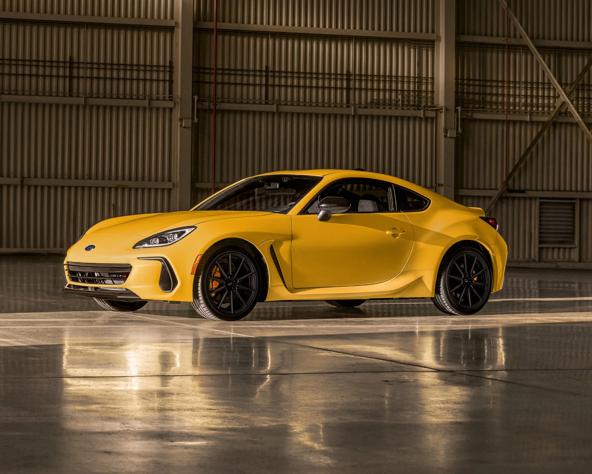 Yellow Subaru BRZ sports car with black wheels, parked indoors on a polished concrete floor, with a corrugated metal wall in the background.