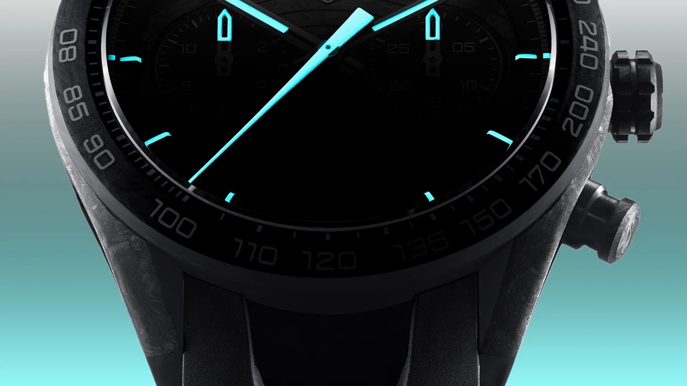 Close-up of a black wristwatch with a tachymeter bezel showing numbers from 80 to 240. The watch hands and hour markers are illuminated in bright blue. The watch has a textured black case and two push buttons on the right side. The background is a gradient from light gray at the top to turquoise at the bottom.