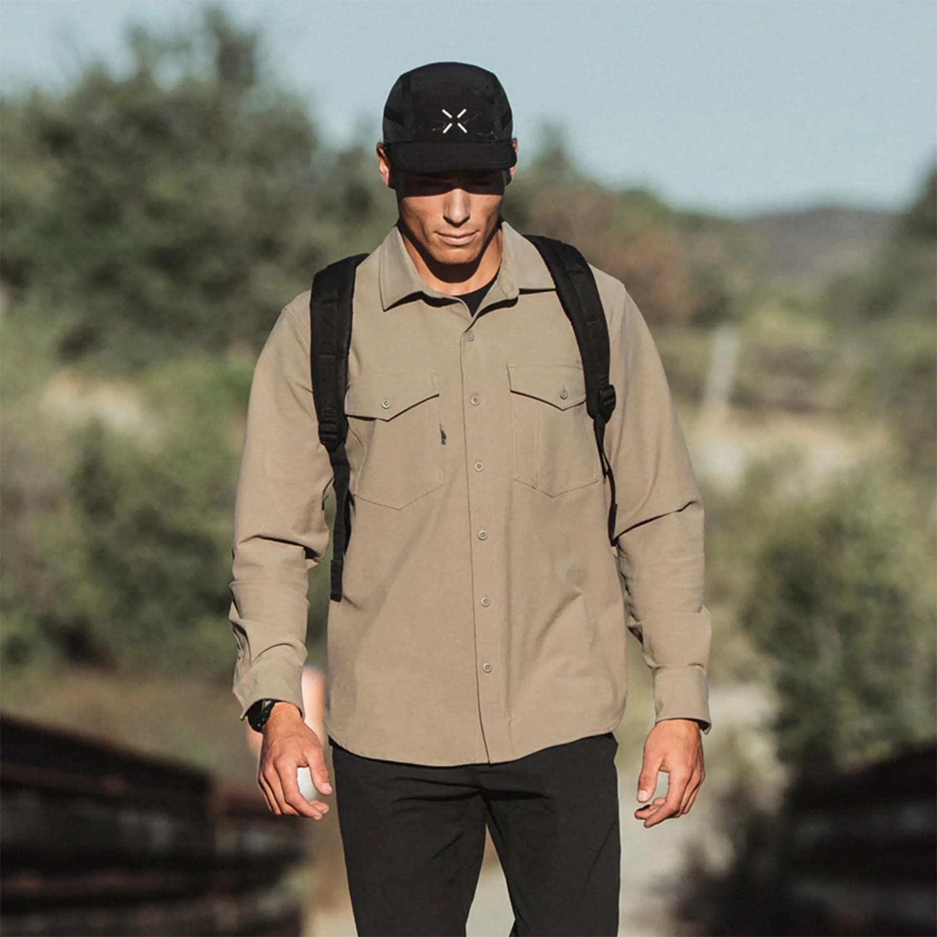 A man wearing a beige button-up shirt with two chest pockets, black pants, a black backpack, and a black cap with a white logo. The background is outdoors with blurred greenery.