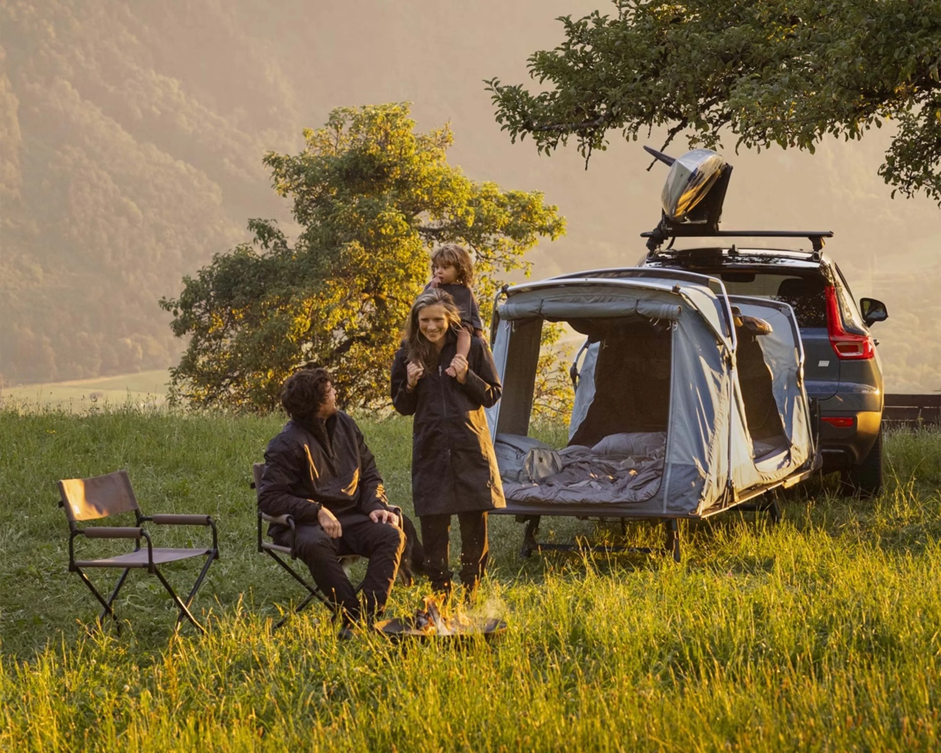 A family camping scene with a man sitting on a folding chair, a woman standing with a child on her shoulders, and a car equipped with a rooftop kayak and an attached tent extending from the rear. The tent is set up on a grassy field near a tree, with a small campfire burning in front of the seated man. The background features a misty, forested hillside bathed in warm, golden light.