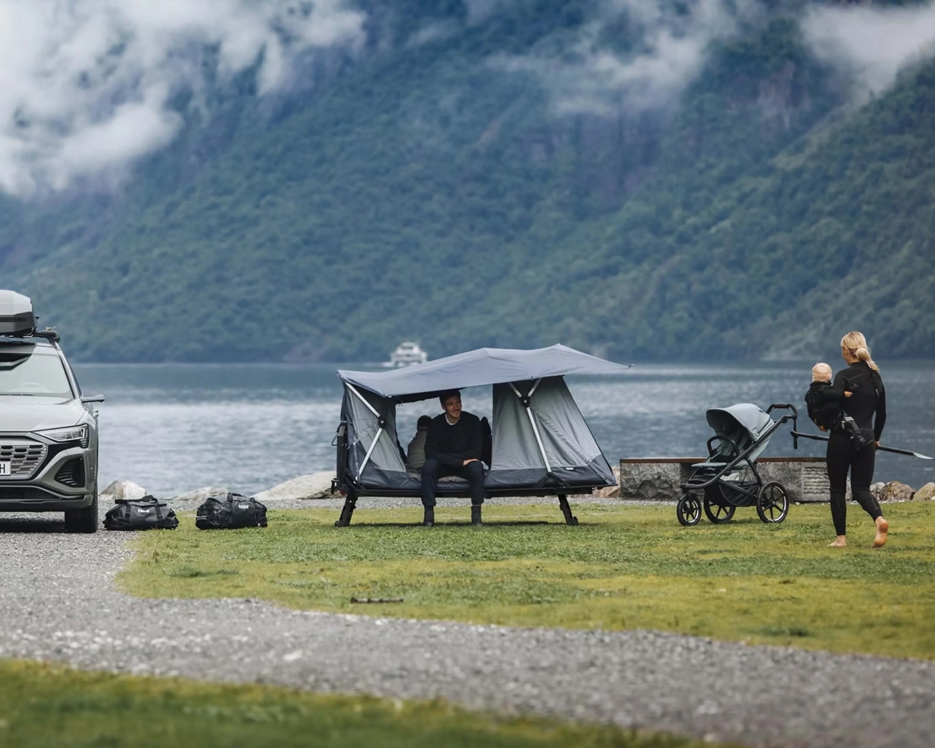 A man sits inside a modern, elevated gray tent with an extended canopy, set up on a grassy area near a lake. To the left, a gray SUV is parked with two black bags on the ground beside it. To the right, a woman dressed in black holds a baby and walks barefoot near a stroller. In the background, there are forested mountains partially covered by clouds.
