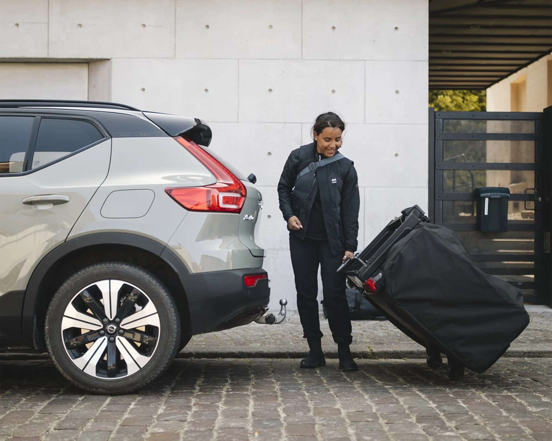 A woman dressed in black is standing next to a silver-gray Volvo XC40, pulling a large black wheeled suitcase. The scene is set on a cobblestone surface in front of a modern concrete wall and a black metal gate. The woman is smiling and looking down at the suitcase.