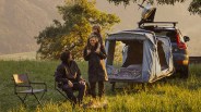 A family camping scene with a man sitting on a folding chair near a small campfire, a woman standing with a child on her shoulders, and a car equipped with a rooftop kayak and an attached tent with bedding, set in a grassy area with trees and hills in the background during golden hour.