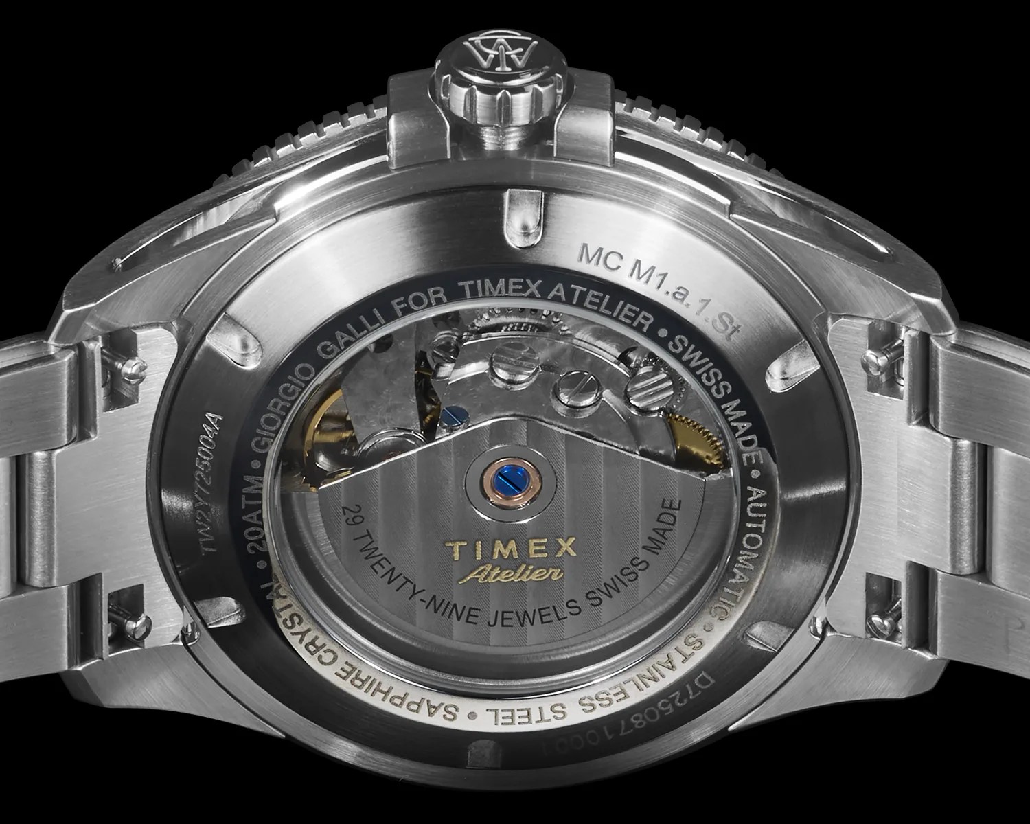 Back view of a stainless steel Timex Atelier wristwatch showing the automatic movement with visible gears and screws. The case back has engravings including "MC M1.a.1 St," "Giorgio Galli for Timex Atelier," "Swiss Made," "Automatic," "Stainless Steel," "Sapphire Crystal," and "29 Twenty-Nine Jewels Swiss Made." The watch has a metal bracelet and a crown with an engraved logo.