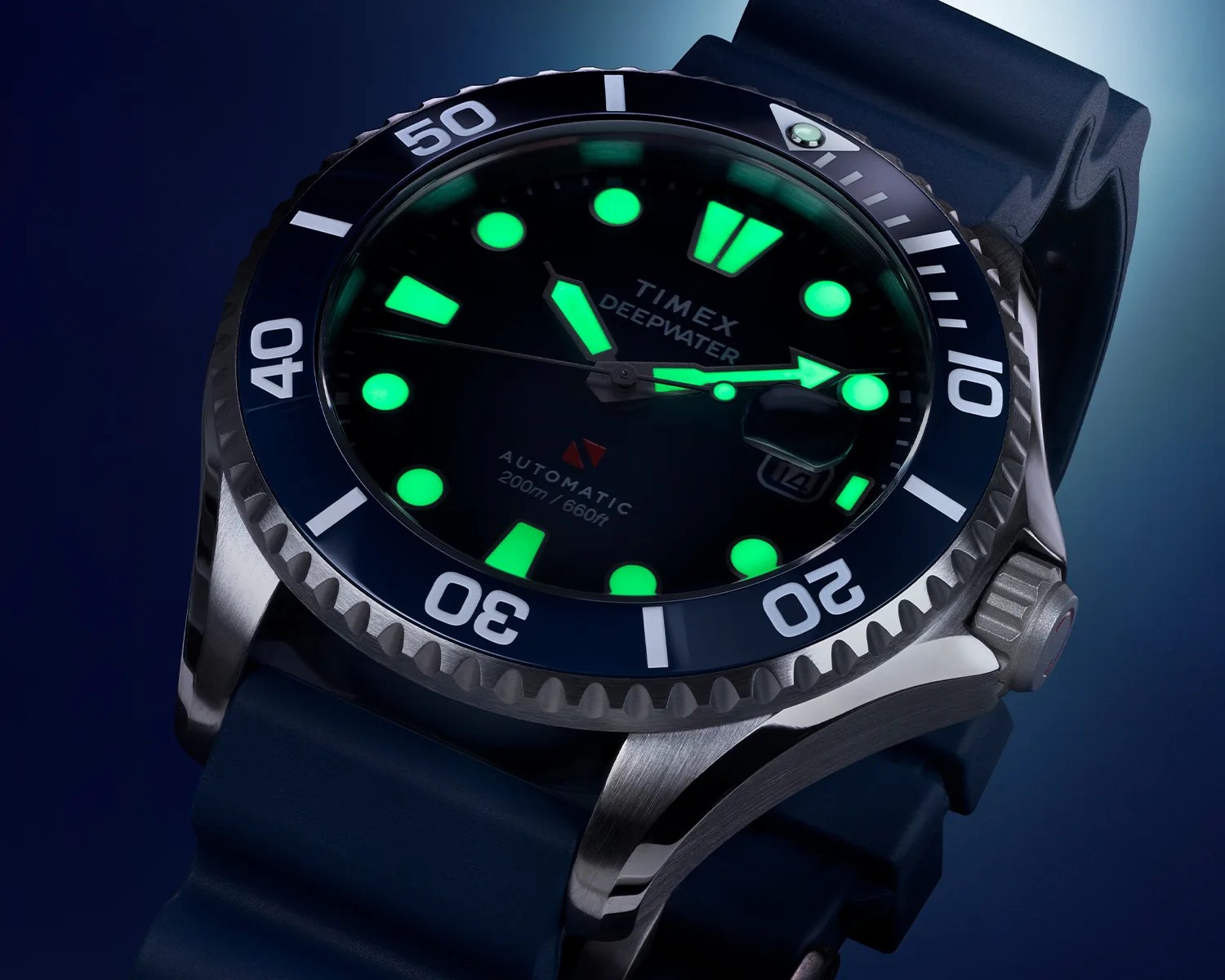 Close-up of a Timex Deepwater automatic dive watch with a stainless steel case, blue bezel marked with white numerals, and a blue rubber strap. The watch face is dark with luminous green hour markers and hands, and a date window at the 3 o'clock position. The bezel has a luminous marker at the 12 o'clock position.
