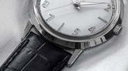 Close-up of a silver wristwatch with a white dial, silver raised numerals, and silver hands. The watch has a polished metal case and a black leather strap with visible stitching. The background is a textured white surface.