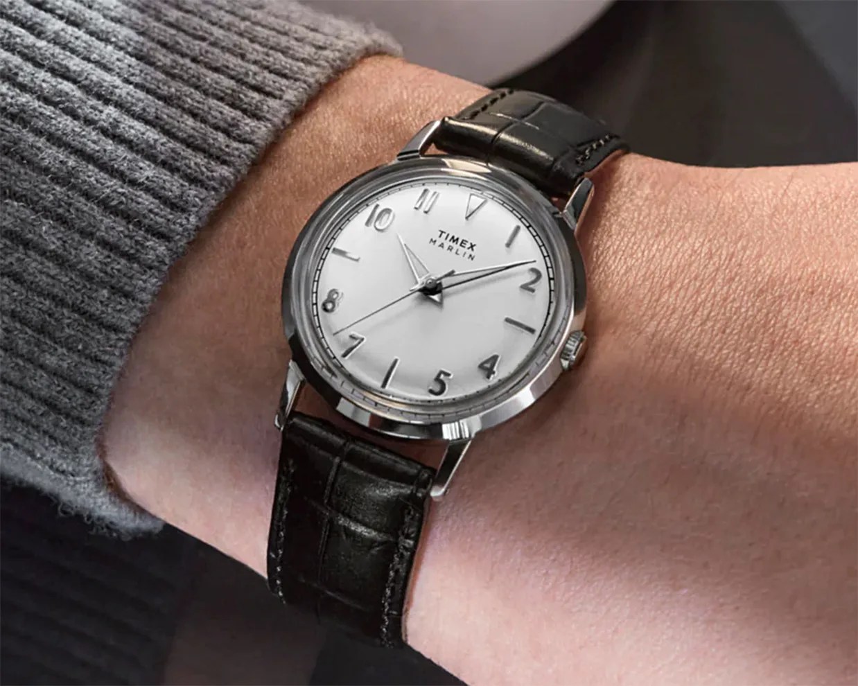 Timex Marlin wristwatch with a white dial, silver hour markers, and black leather strap, worn on a wrist with a gray knitted sleeve visible.