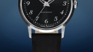 Close-up of a wristwatch with a black dial featuring silver Arabic numerals and hands. The watch face includes the text "WATER RESISTANT" and "TODD SNYDER" near the bottom. The watch has a polished silver bezel and a black leather strap with visible stitching. The background is a gradient of dark blue.