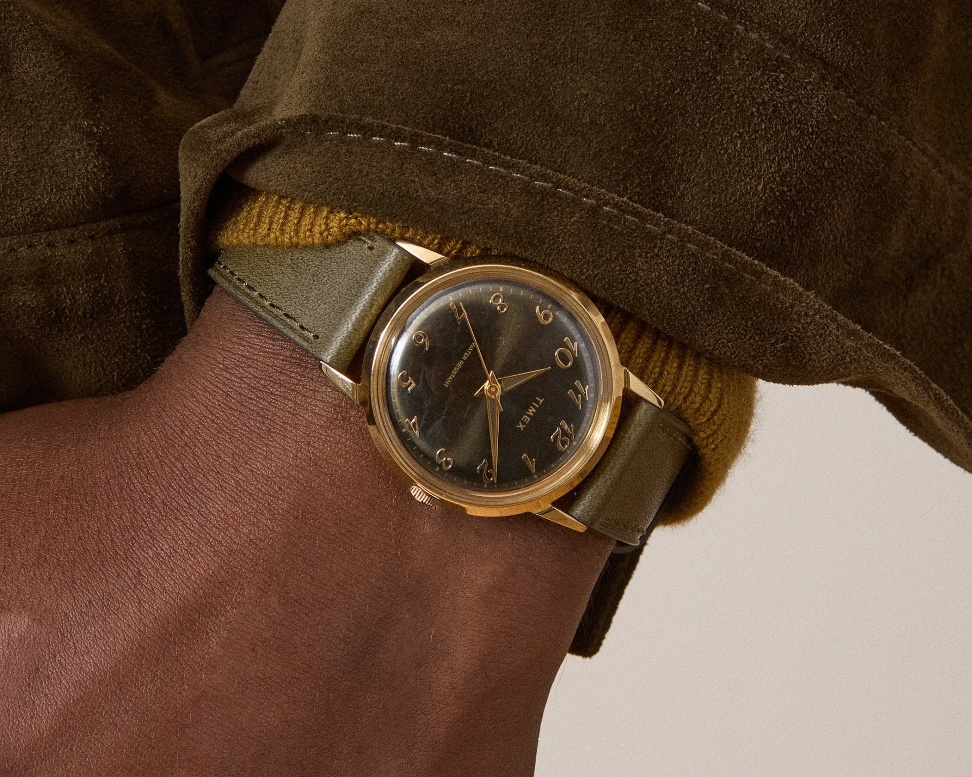 A wristwatch with a gold-tone case and a black dial featuring gold Arabic numerals and hands. The watch has a green leather strap and is worn on a wrist with dark skin. The wearer is dressed in a brown suede jacket and a mustard yellow knit sweater.