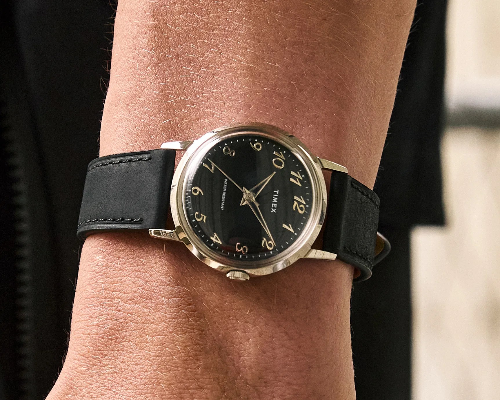 Silver Timex wristwatch with a black dial and gold Arabic numerals, featuring a black leather strap, worn on a wrist.
