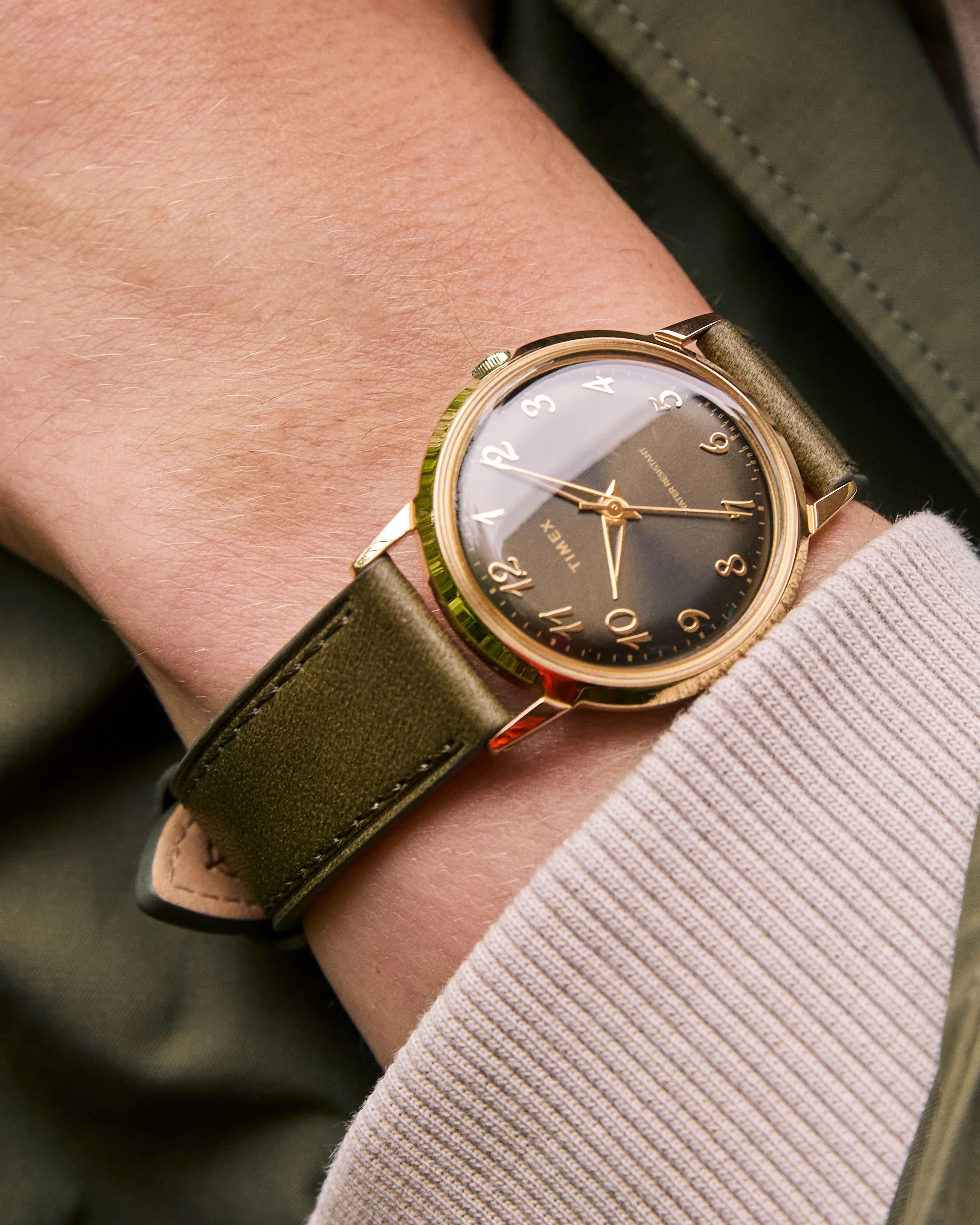 Gold-toned Timex wristwatch with a dark brown dial featuring gold Arabic numerals and hands, paired with an olive green leather strap, worn on a wrist with a beige ribbed sleeve and an olive green jacket.