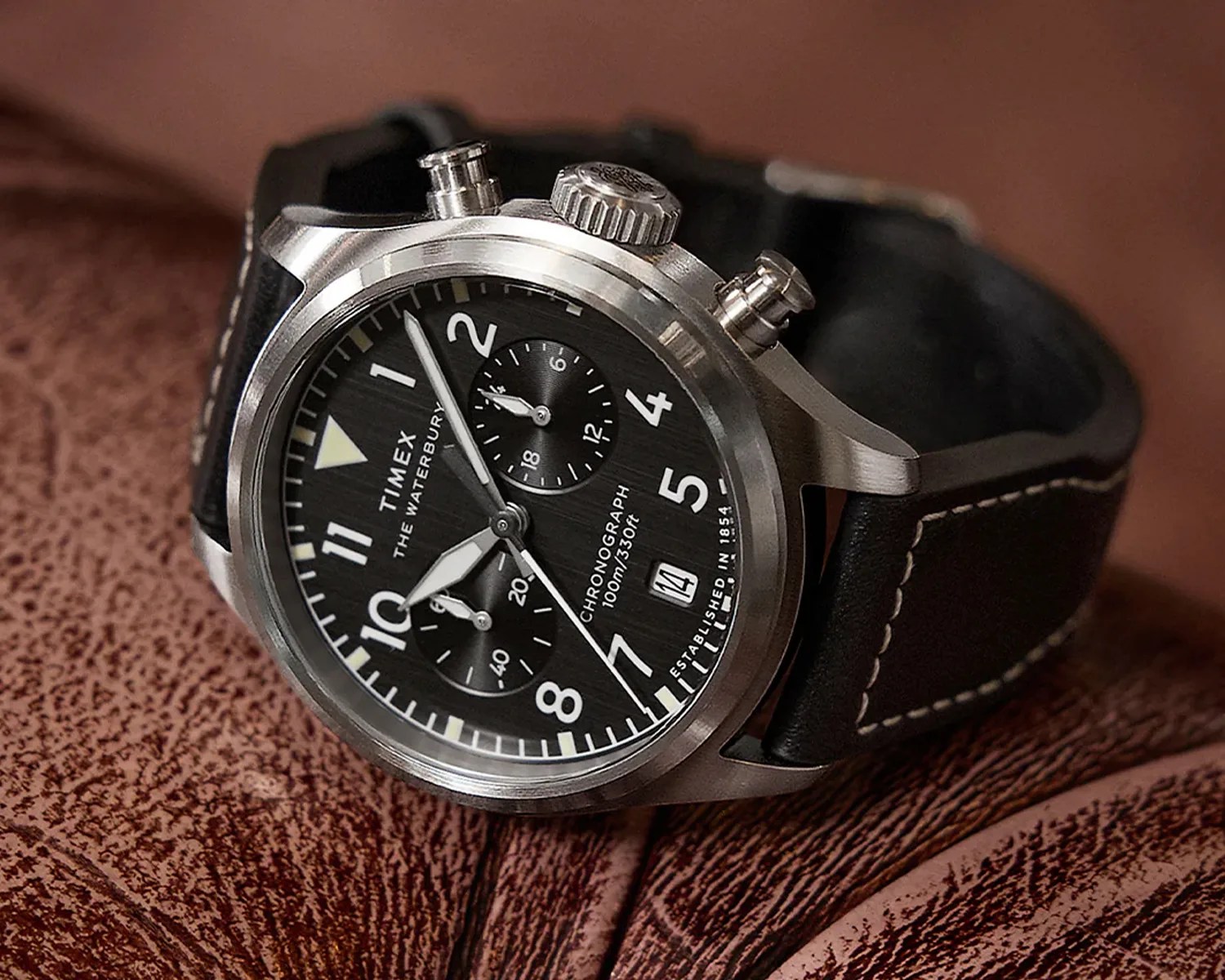 Timex chronograph wristwatch with a black dial, white numerals, and two subdials. The watch has a stainless steel case and a black leather strap with white stitching. The dial includes a date window at the 4 o'clock position and text indicating "THE WATERBURY," "CHRONOGRAPH 100m/330ft," and "ESTABLISHED IN 1854." The watch is resting on a textured brown leather surface.