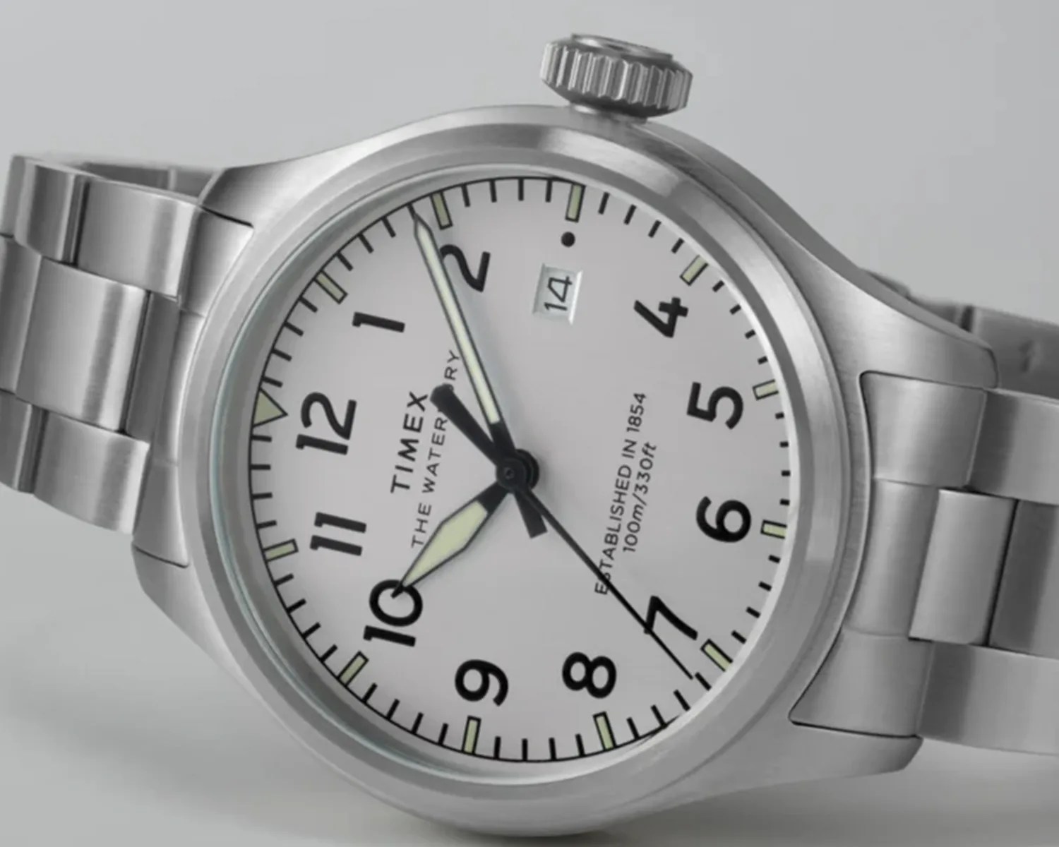 Silver Timex wristwatch with a white dial, black numerals, and luminous hands. The watch features a date window at the 3 o'clock position and a stainless steel link bracelet. The dial also includes the text "TIMEX THE WATERBURY" and "ESTABLISHED IN 1854 100m/330ft.