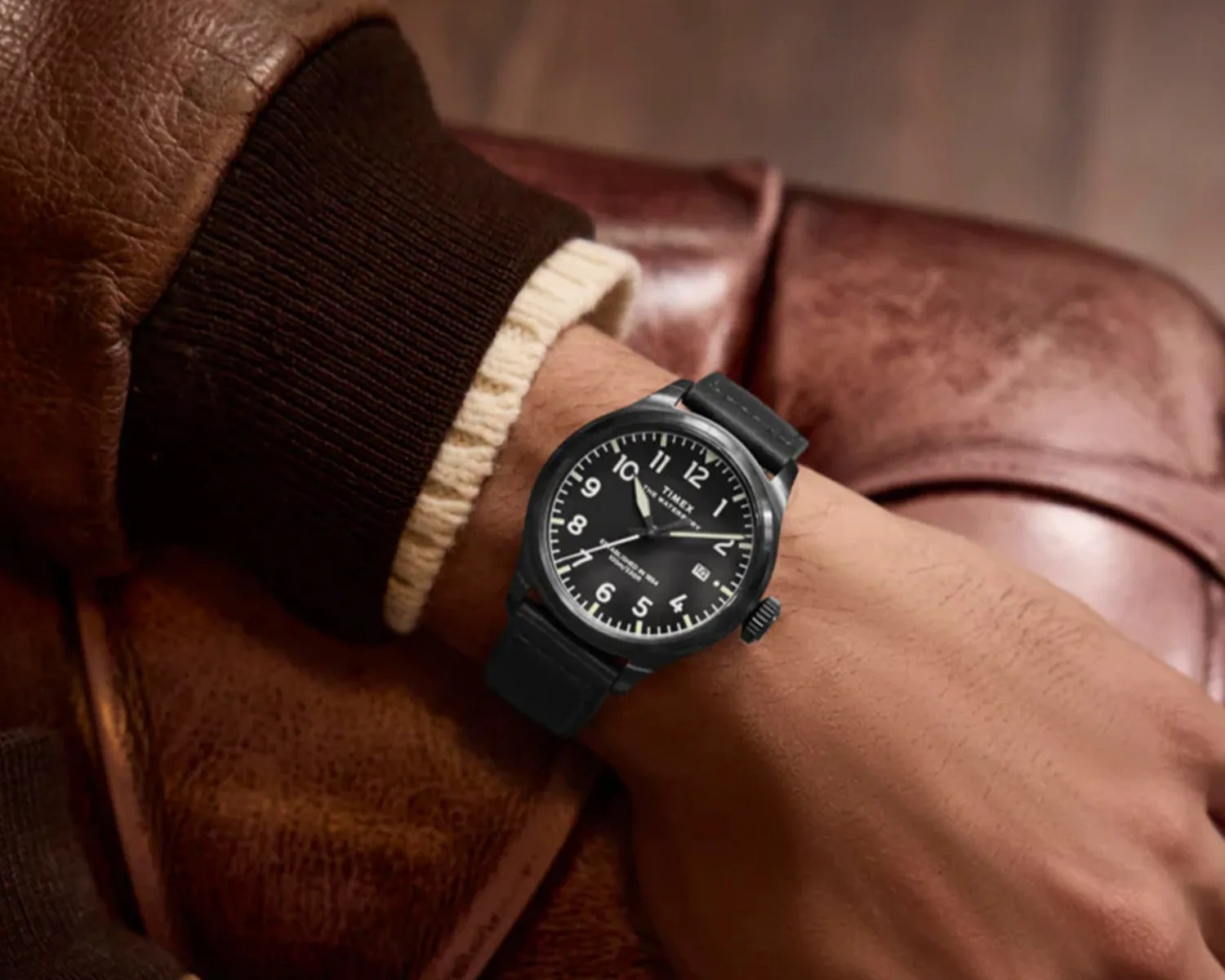 Black Timex wristwatch with a black leather strap, white numerals, and a date display, worn on a wrist with a brown leather jacket and cream-colored sweater sleeve visible.