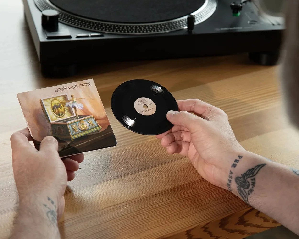 Hands holding a miniature vinyl record and its cover titled "Rainbow Kitten Surprise" with an image of a ballerina on a music box, placed on a wooden table near a turntable.