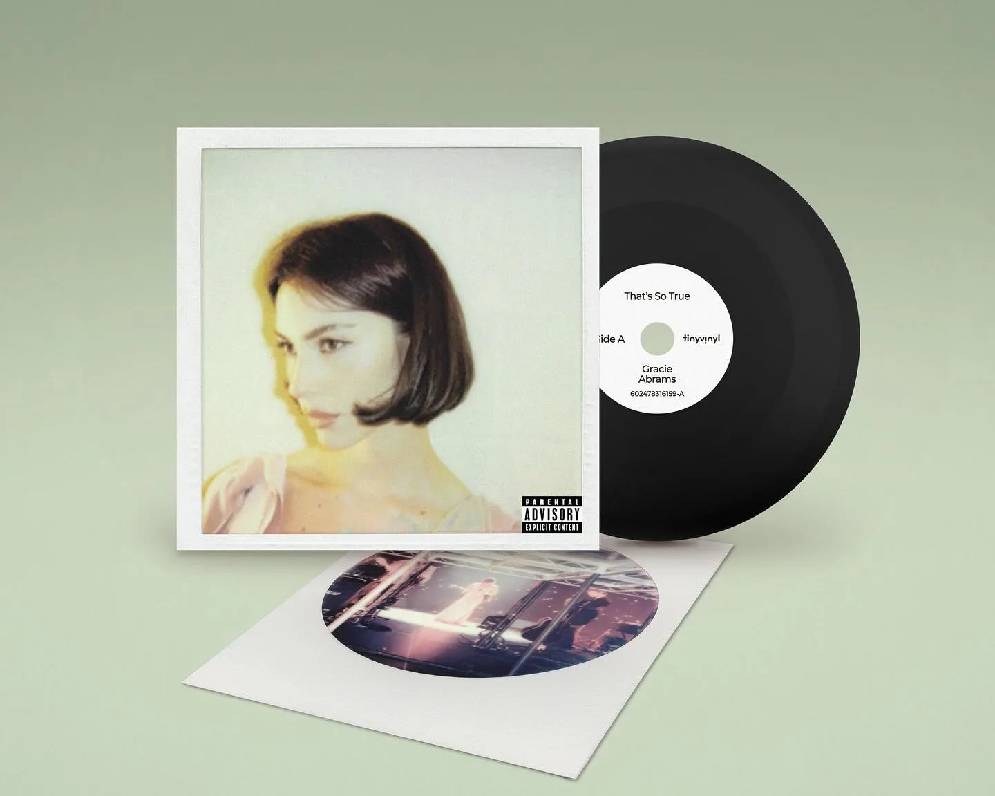 A vinyl record with a black disc and a white label that reads "That's So True," "Side A," "Gracie Abrams," and "tinyvinyl." The record is partially inside a square album cover featuring a soft-focus portrait of a woman with short dark hair looking to the side. The cover has a parental advisory explicit content label in the bottom right corner. Below the record and cover is a white sleeve with a circular photo of a stage performance. The background is a muted green.