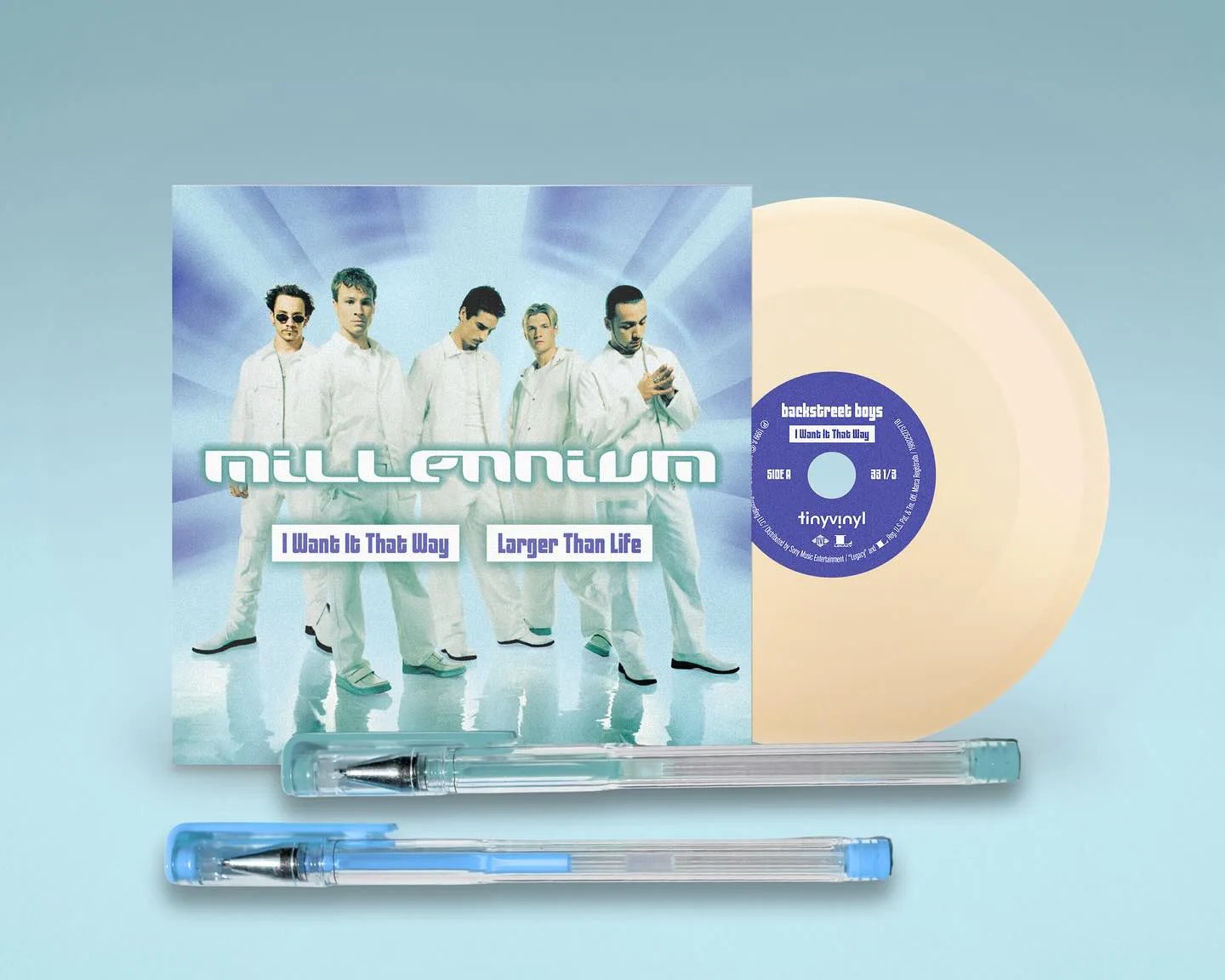 A cream-colored vinyl record partially inside a cover featuring the Backstreet Boys dressed in white with a blue background. The cover has the word "millennium" in large white letters and the song titles "I Want It That Way" and "Larger Than Life." Two ballpoint pens, one with a blue cap and one with a clear cap, are placed below the record and cover for size comparison.