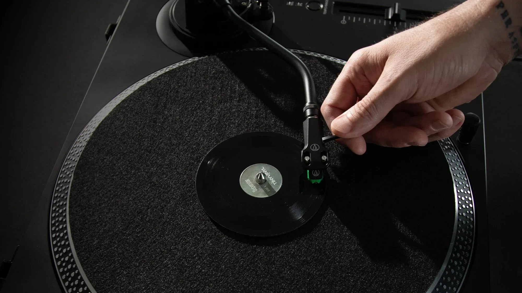 A close-up of a hand adjusting the tonearm of a black turntable with a small vinyl record labeled "Superstar" and "TINYMUZ" on the platter. The turntable has a textured black mat and a silver dotted edge.