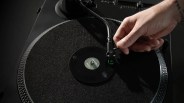 A close-up of a hand adjusting the tonearm of a black turntable with a small vinyl record labeled "Superstar" and "TINYMUZ" on the platter. The turntable has a textured black mat and a silver dotted edge.