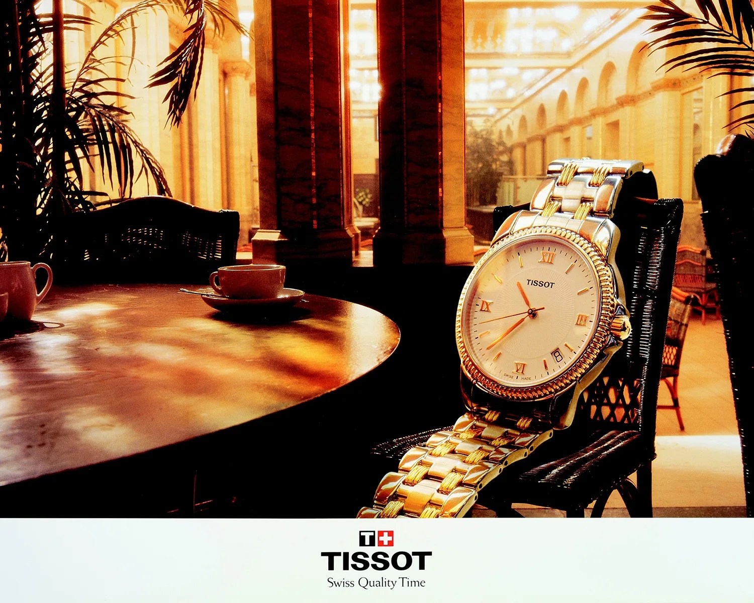 A gold and silver Tissot wristwatch with a white face and Roman numeral hour markers is displayed on a black wicker chair next to a round wooden table with a cup and saucer. The background features an elegant, warmly lit interior with tall columns and arched windows. The bottom of the image shows the Tissot logo and the tagline "Swiss Quality Time.