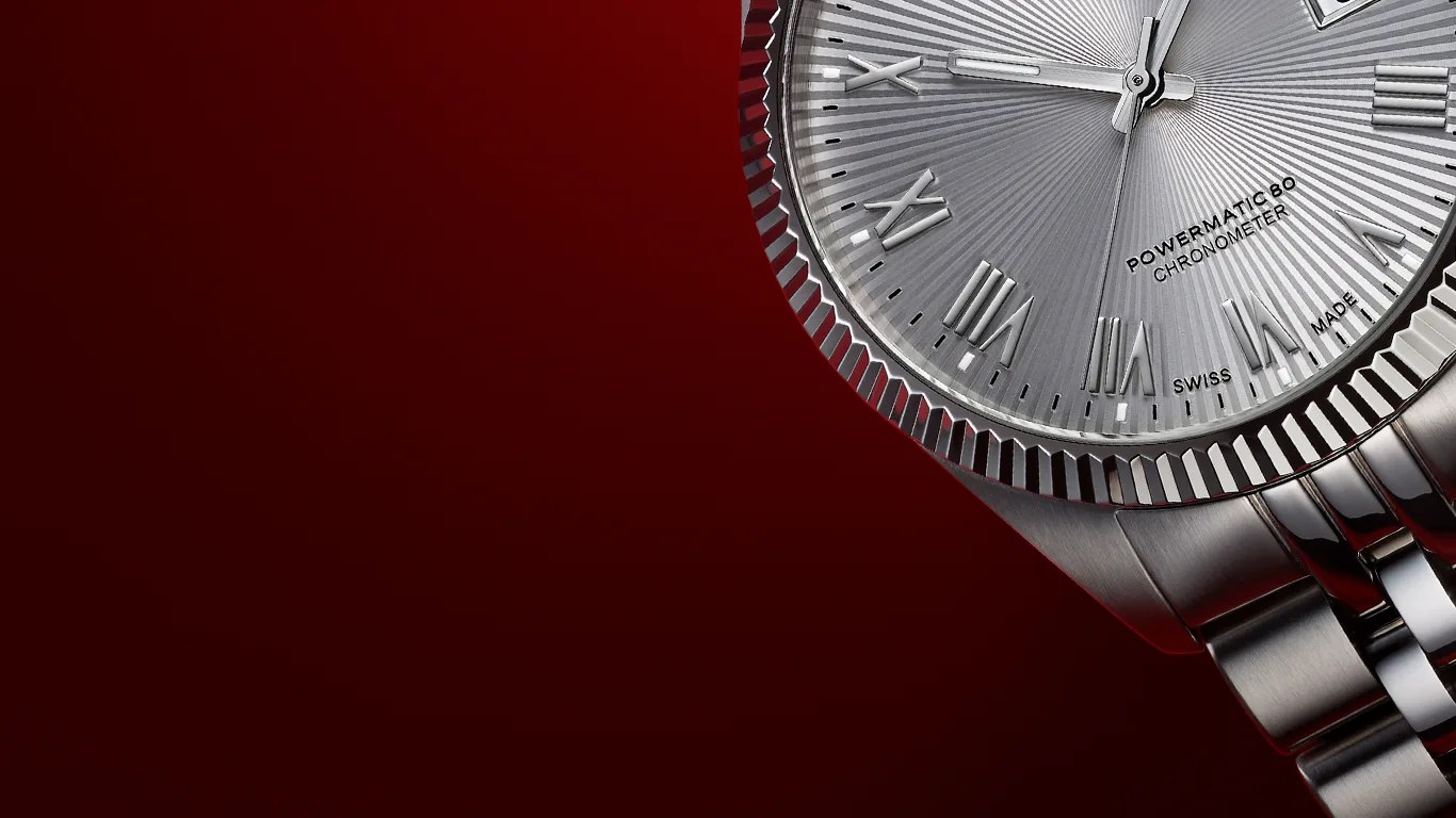 Close-up of a silver Tissot wristwatch with a fluted bezel and a metal link bracelet. The watch face features Roman numeral hour markers, a textured radial pattern, and the text "POWERMATIC 80 CHRONOMETER" and "SWISS MADE." The background is a deep red gradient.