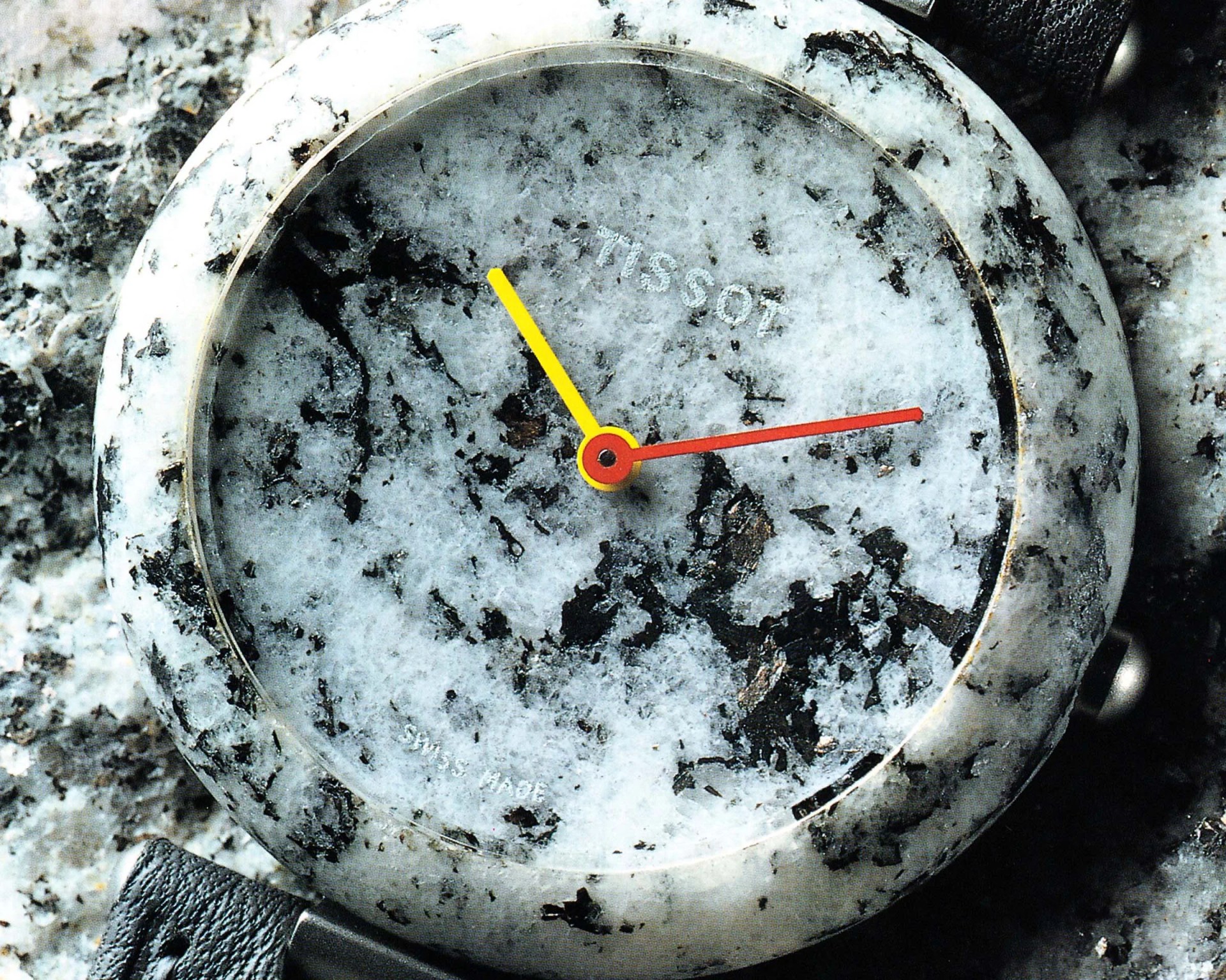 A close-up of a Tissot wristwatch with a unique marble-like textured face and case in shades of white and black. The watch has a yellow hour hand, a red minute hand, and no visible numbers or markers on the dial. The strap appears to be black leather. The words "TISSOT" and "SWISS MADE" are faintly visible on the watch face.