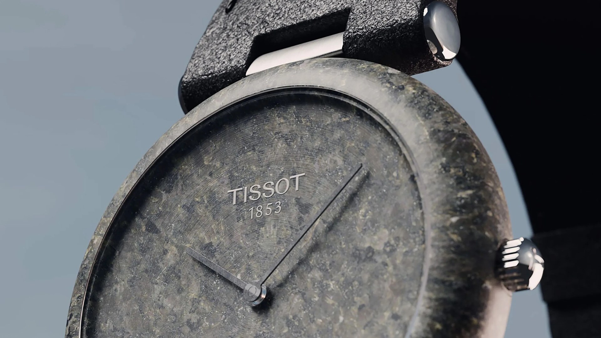 Close-up of a Tissot wristwatch with a textured, stone-like gray dial and case. The watch has minimalist design with thin, dark hands and the brand name "TISSOT 1853" displayed near the center. The strap appears to be black with a matte finish, and the crown is metallic and shiny.