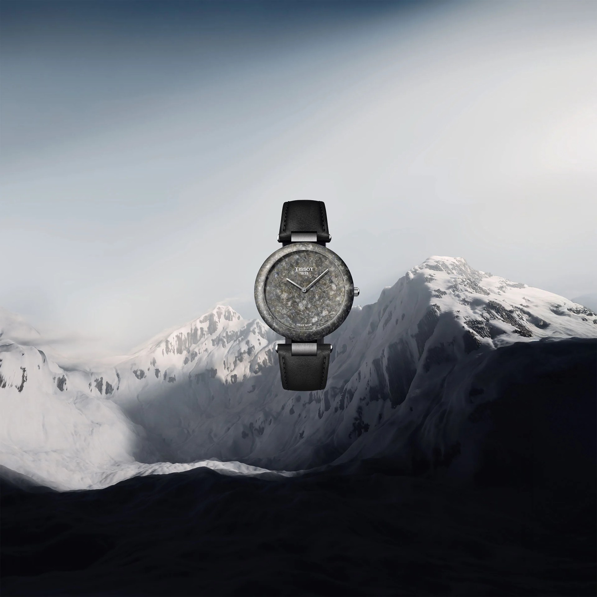 Round wristwatch with a textured stone-like gray dial and bezel, featuring silver hour and minute hands, and a black leather strap, set against a backdrop of snow-covered mountain peaks under a cloudy sky. The watch brand "Tissot 1853" is visible on the dial.