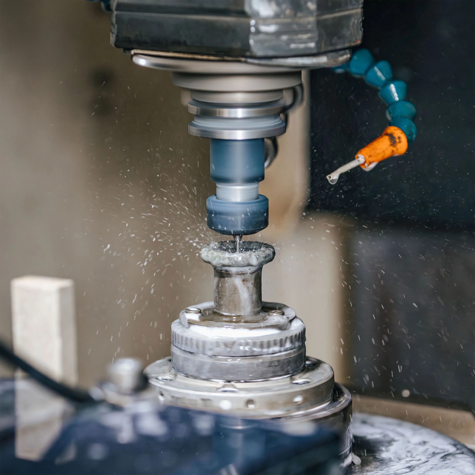 A close-up of a CNC milling machine cutting a metal workpiece, with coolant fluid spraying around the cutting area. The machine's spindle and cutting tool are visible, along with a flexible coolant hose with an orange nozzle.