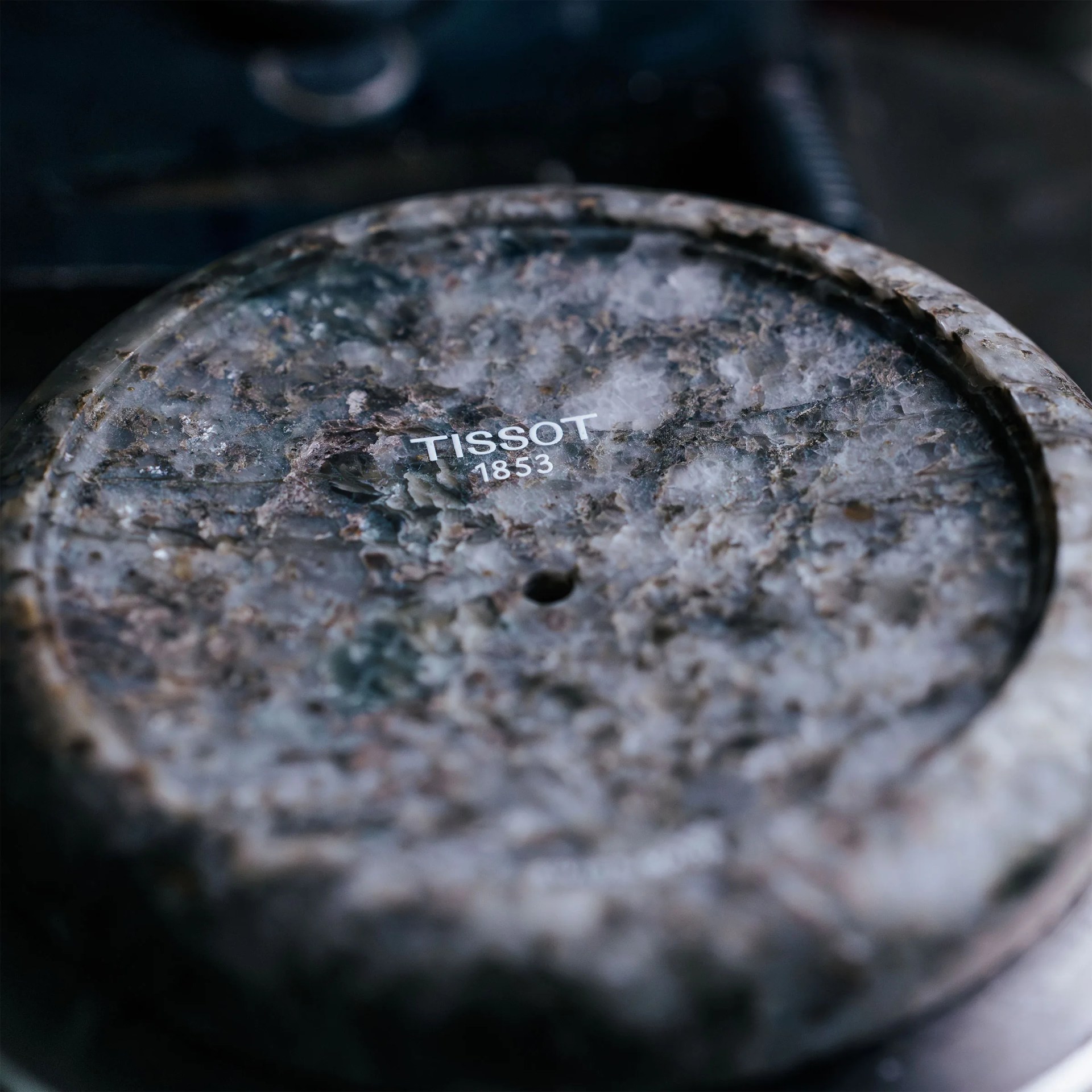 Close-up of a textured, round stone surface with the word "TISSOT" and the year "1853" printed in white on it. The surface has a rough, marbled appearance with shades of gray, brown, and white.