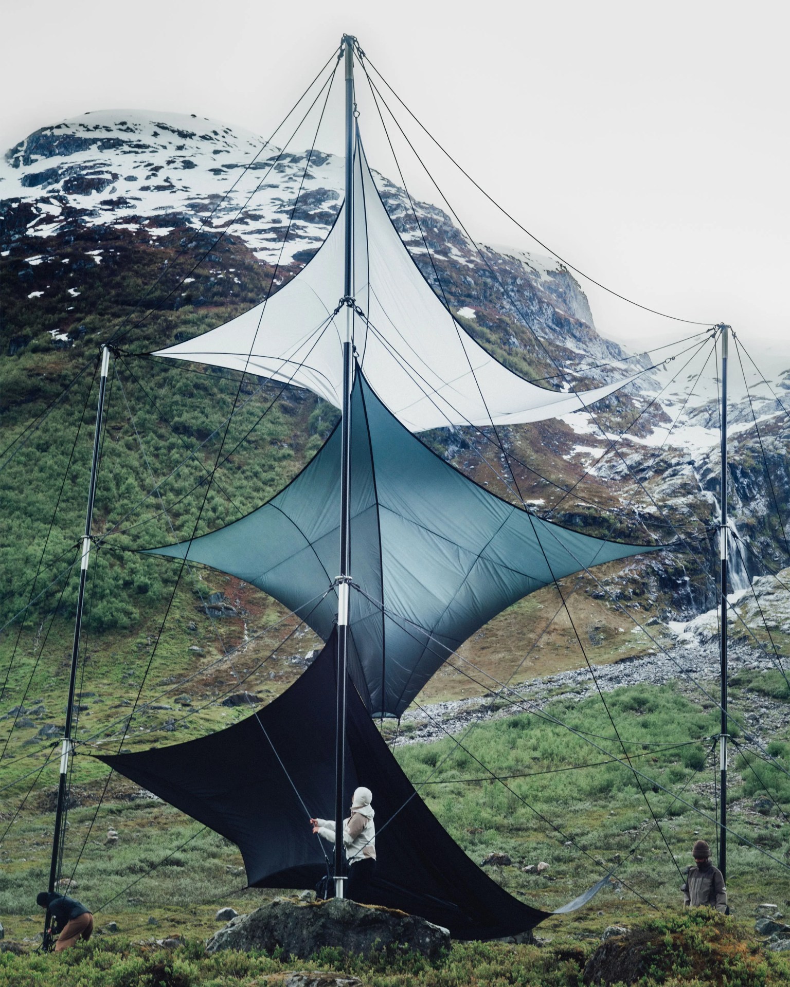 A large, multi-tiered fabric structure supported by tall poles and numerous tensioned cables, set against a mountainous landscape with patches of snow and greenery. Three people are adjusting the cables and fabric, which consists of three differently colored sections: white at the top, gray in the middle, and black at the bottom. The structure appears to be an artistic installation or a specialized shelter.
