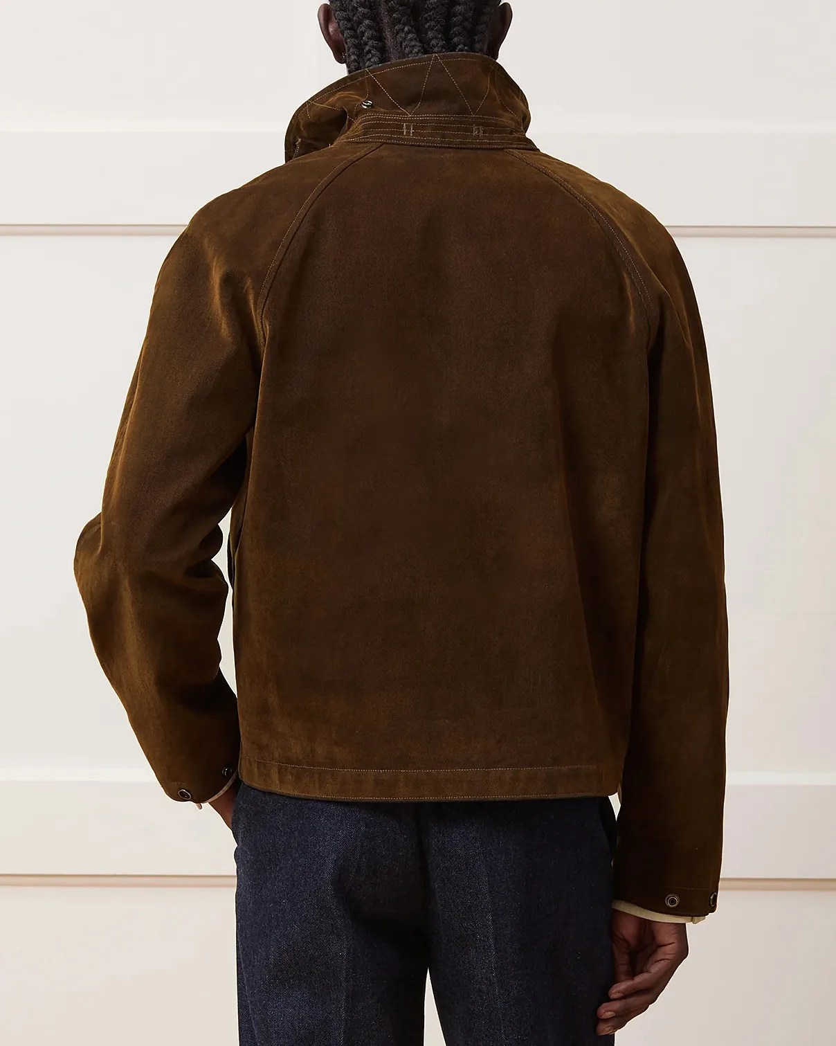 Dark brown suede jacket with a high collar and visible stitching details, shown from the back. The jacket has long sleeves with snap button cuffs and a straight hem. The person wearing it has dark braided hair and is also wearing dark blue pants.
