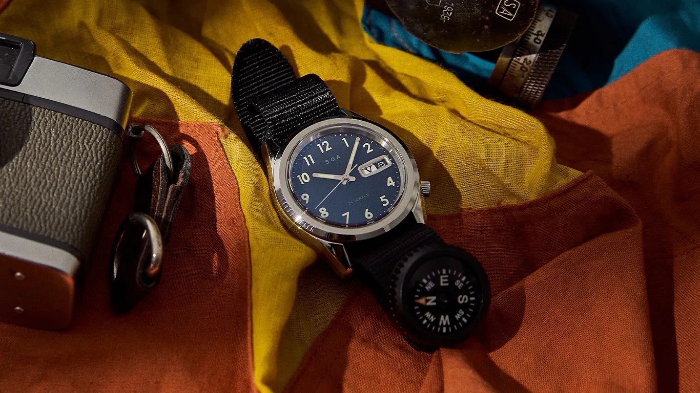 A wristwatch with a black fabric strap featuring a built-in compass. The watch has a silver bezel, a dark blue dial with white numerals, and a date display showing "V 8". It is placed on a multicolored fabric background with yellow, orange, and blue sections. Part of a vintage camera is visible on the left side of the image.