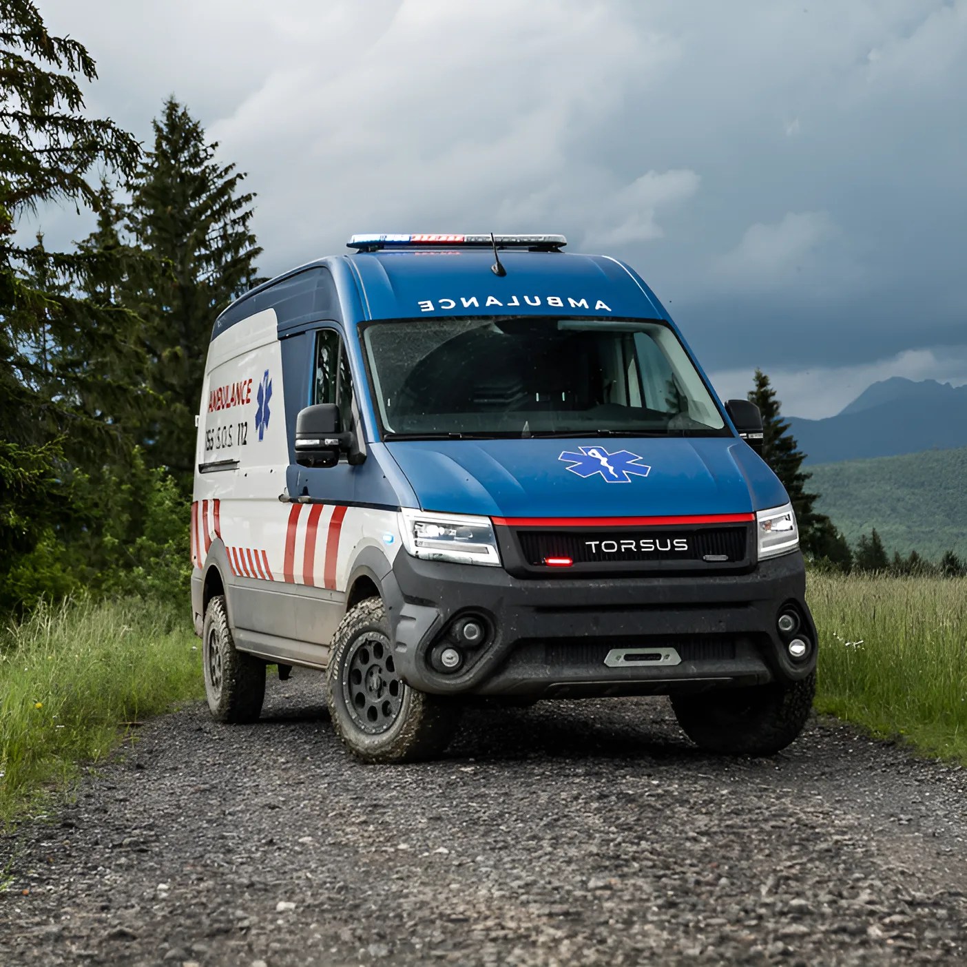 Off-road ambulance van with blue and white body, red stripes on the side, and "TORSUS" branding on the front grille, parked on a gravel path surrounded by grass and trees under a cloudy sky.