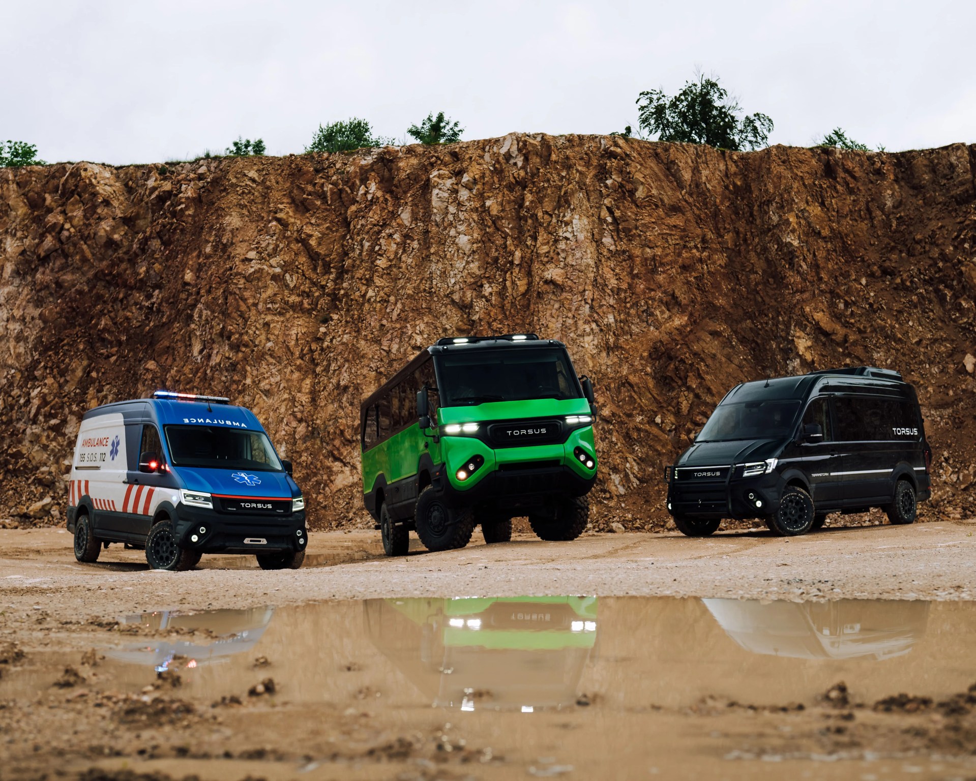 Three off-road vehicles are parked on a dirt surface in front of a rocky cliff. From left to right, there is a blue and white ambulance van with red stripes and emergency lights, a green and black rugged bus with large tires, and a black van with tinted windows. All vehicles have the brand name "TORSUS" displayed on the front. The ground in front of the vehicles has a large puddle reflecting their images.