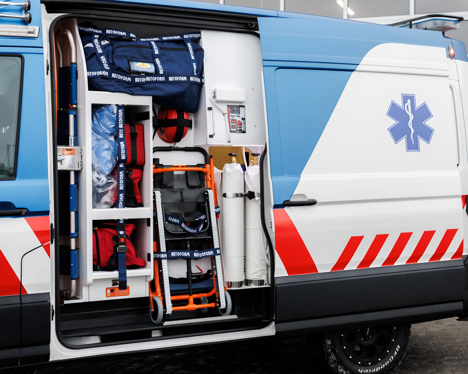 Side view of an ambulance with its sliding door open, revealing organized medical equipment inside, including oxygen tanks, a foldable stretcher, red and blue medical bags, and other emergency supplies secured with straps. The exterior of the ambulance is white with blue and red markings and features a blue Star of Life symbol.