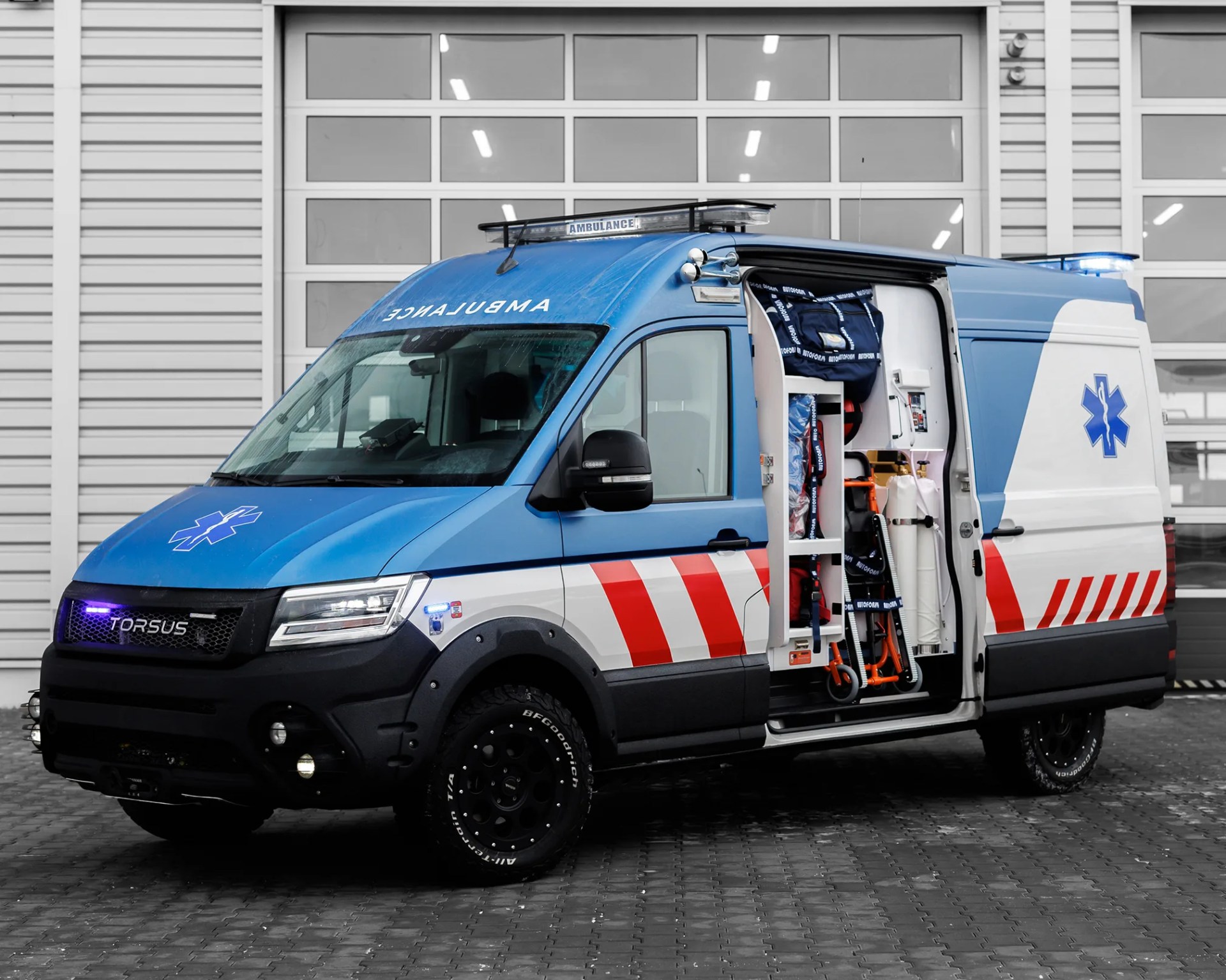 A blue and white off-road ambulance with red stripes is driving on a dirt path, kicking up dust behind it. The vehicle has rugged tires and a roof-mounted light bar, with "TORSUS" displayed on the front grille and "AMBULANCE" written on the side along with emergency numbers. 
