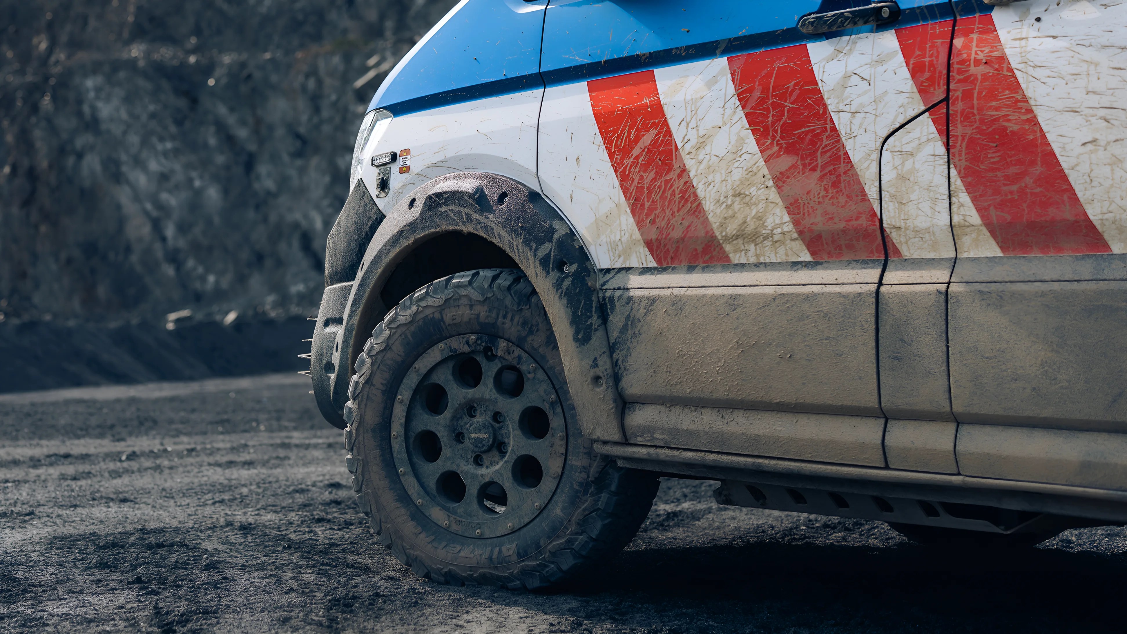 Close-up of the front left side of a muddy off-road vehicle with a blue, white, and red striped paint job, featuring a rugged tire and a black fender flare. The vehicle is parked on a dirt surface with a rocky background.
