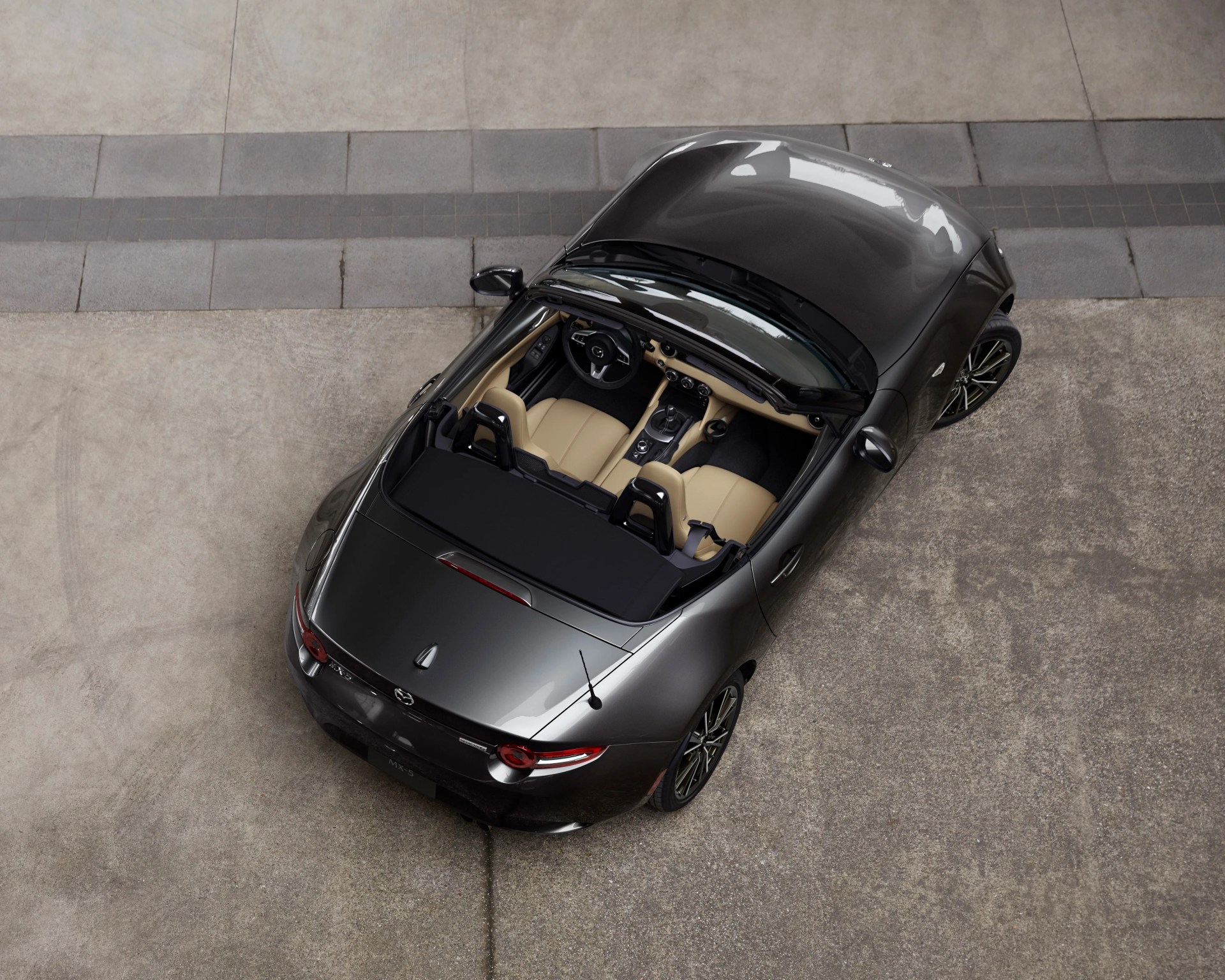 A dark gray convertible sports car with a beige interior, viewed from above. The car has two seats, a black soft top folded down, and sleek, modern design lines. The vehicle is parked on a concrete surface with a tiled strip in the background.