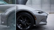 Front side view of a gray Mazda car with water droplets on its surface, featuring a black alloy wheel and a sleek headlight design. The image is partially obscured by a glass panel on the left side.