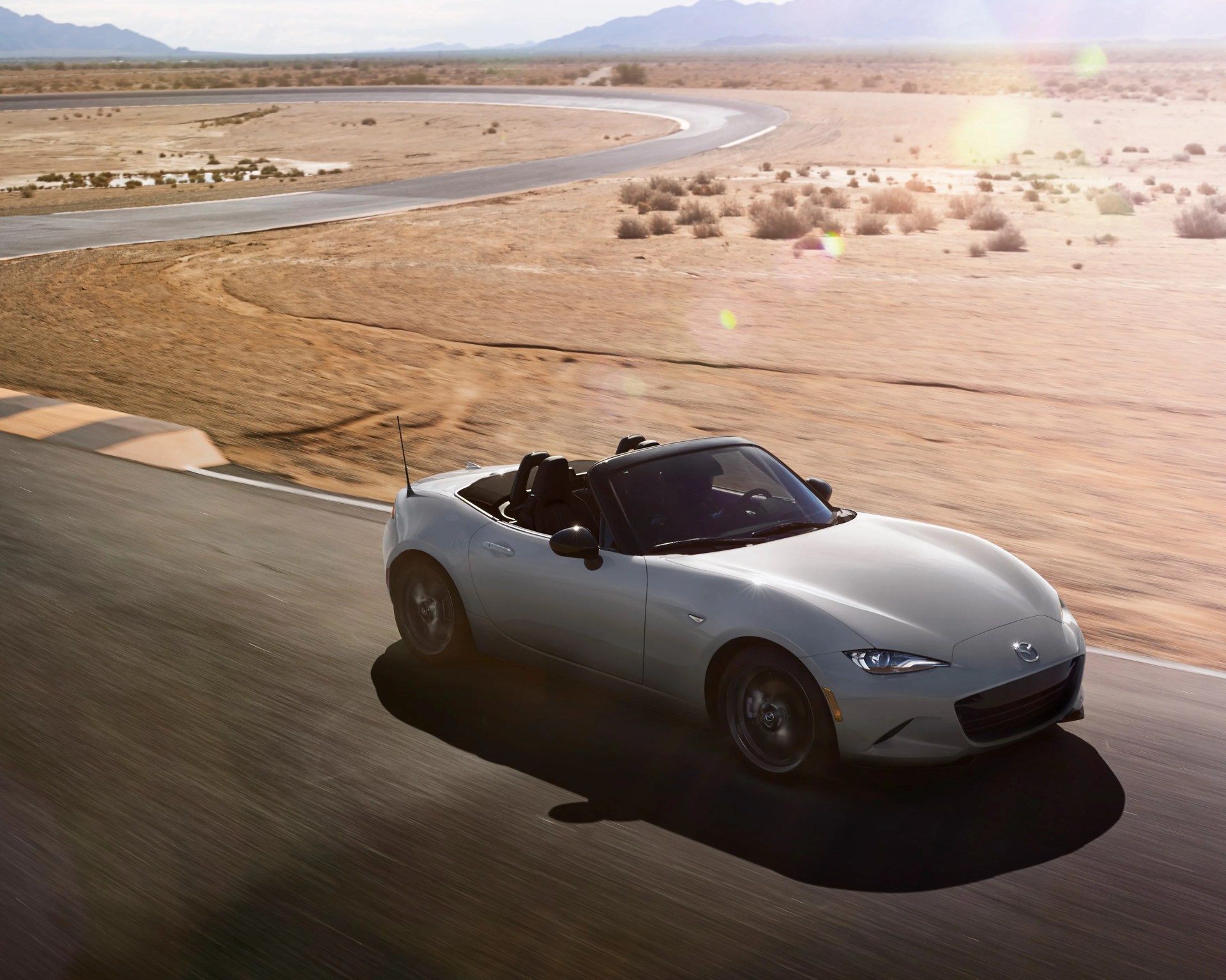 Silver convertible sports car driving on a curved desert racetrack with dry shrubs and mountains in the background under bright sunlight.