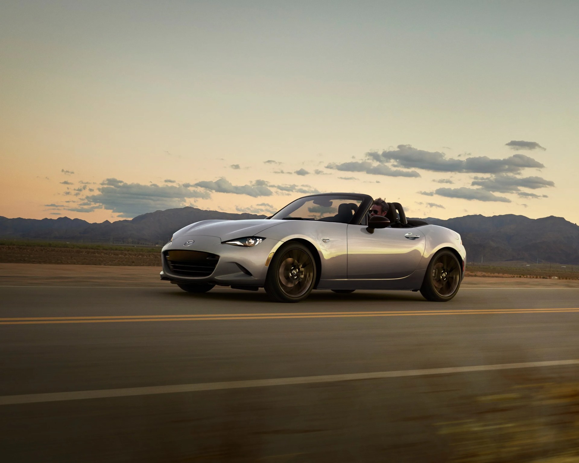 Silver convertible sports car driving on a road at sunset with mountains and a partly cloudy sky in the background.